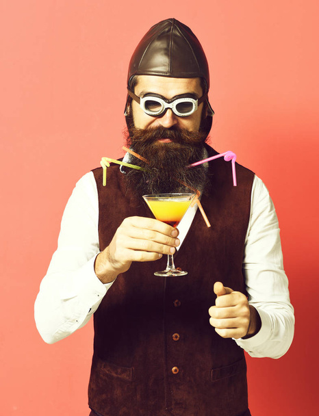 handsome bearded pilot or aviator man with long beard and mustache on serious face holding glass of alcoholic beverage in vintage suede leather waistcoat with hat and glasses on red studio background - Foto, imagen