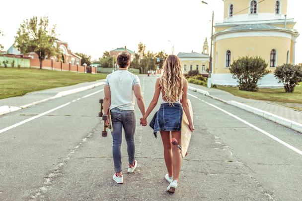若いカップルは、スケートボード、ロングボードの手で、市内の夏に歩きます。バックビュー。週末の散歩、若者のライフスタイル. - 写真・画像