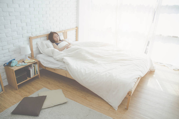 Chica asiática duerme en pijamas grises y pantalones cortos. En la cama en el dormitorio blanco.Las manchas oscuras en la cara de una joven causada por la luz del sol.Algodón y soleado
. - Foto, imagen