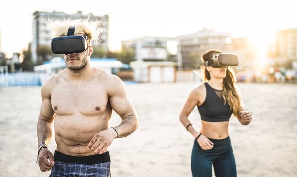 Jeune couple courant à la plage avec casque de lunettes de réalité virtuelle - Concept sport et technologie avec des amis jogging et exercice sur la simulation de trek en été - Filtre de coucher de soleil contrasté
 - Photo, image