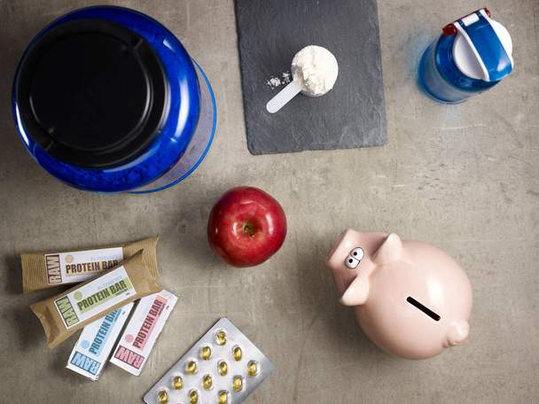 Closeup on big blue protein jar, shaker, measuring spoon with powder, raw protein bars, red apple, sport supplements in tablets and piggy bank laying on the floor. - Foto, Bild