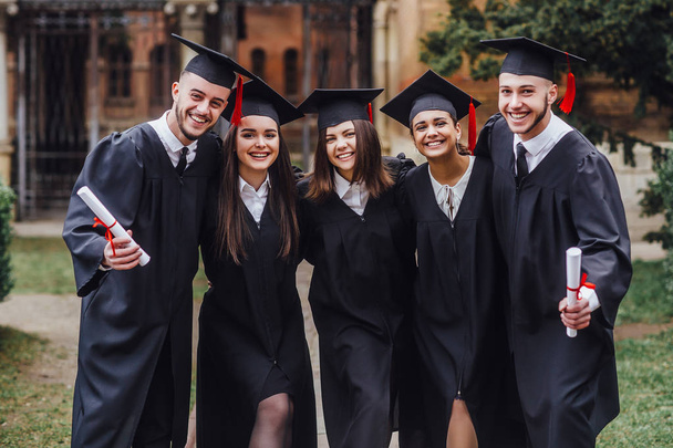 Absolwenci szkół wyższych w sukien dyplomowych stojących blisko siebie i uśmiechając - Zdjęcie, obraz