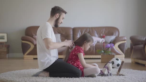 Young bearded father brushing the hair of his little girl while the child combing the tail of her toy horse sitting on the floor on fluffy carpet. Family leisure. Fatherhood, caring, love. Side view - Video, Çekim
