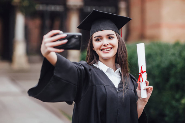 Graduated woman in graduation hat and gown making selfie - Фото, изображение