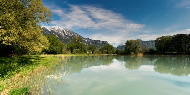 весенний пейзаж с заснеженными горами и прудом в зеленом
 - Фото, изображение