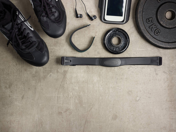 Closeup on laying on the floor black sneakers, headphones, fitness tracker, hear rate monitor, smartphone in running armband, bottle of water, weight plates. - Photo, Image