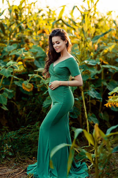 Pregnant european woman in a field of sunflowers, beautiful young european woman waiting for a child, prenant woman with dark hair in long green dress on the nature - Foto, afbeelding