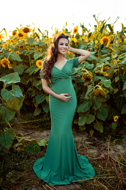 Pregnant european woman in a field of sunflowers, beautiful young european woman waiting for a child, prenant woman with dark hair in long green dress is smiling on the nature - Foto, afbeelding