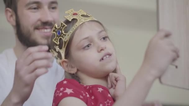 Portrait attractive young bearded father putting the crown on the head of his little girl, making her princess while the child looking in the mirror close up. Family leisure. Fatherhood, caring, love - Video