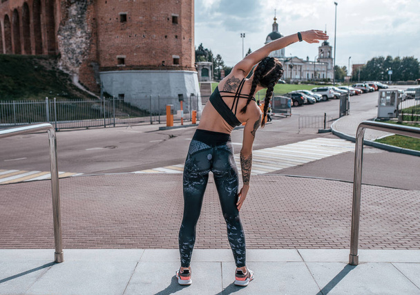 The girl in tattoos, in the summer in city, trains fitness, gymnastics workout. Sportswear leggings top. In the morning before the run. Back view. Background of the road, valet parking, buildings. - Photo, Image