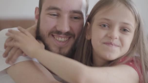 Close up portrait handsome bearded man and cute positive girl looking in the camera smiling, the child hugging the man. Father and daughter having fun at home, leisure of the happy family - Footage, Video
