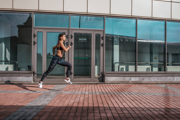 A beautiful woman, a girl runs in the summer in city, on a morning jog, tanned skin. Sportswear leggings top. Phone earphones. Fitness concept jogging, lifestyle. Free space for text. - Foto, Imagem