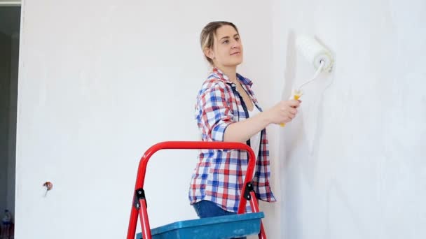 Video of beautiful young woman in checkerd shirt doing renovation at her house. Young girl painting walls with paint roller - Footage, Video