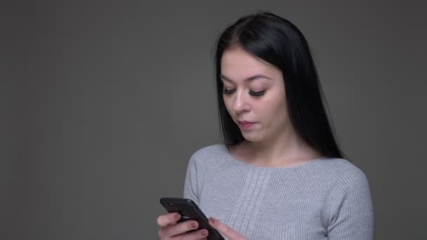 Closeup shoot of young pretty brunette caucasian female texting on the phone with background isolated on gray - Imágenes, Vídeo
