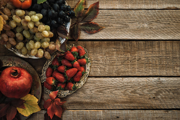 Verse granaatappels, druiven en bessen in een oude vintage plaat met bladeren. Op rustieke houten achtergrond. Vrije ruimte voor tekst. Bovenaanzicht. Getinte afbeelding. - Foto, afbeelding