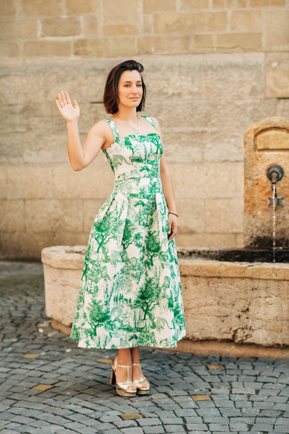 Outdoor fashion portrait of beautiful woman with dark hair, wearing long vintage styled green dress, posing on the city street - Photo, image