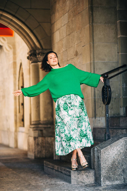 Outdoor fashion portrait of beautiful woman with dark hair, wearing long vintage styled green skirt and pullover, posing on the city street - Foto, Imagen