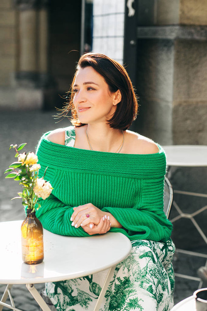 Retrato de moda al aire libre de una hermosa mujer con cabello oscuro, usando jersey verde, posando en la cafetería de la calle
 - Foto, Imagen