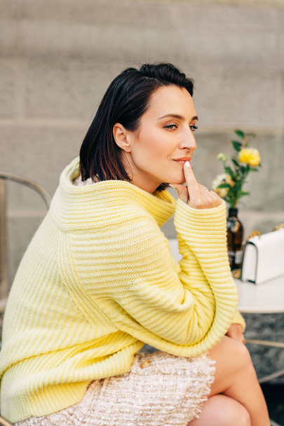 Outdoor fashion portrait of beautiful woman with dark hair, wearing yellow pullover, posing in street cafe - 写真・画像