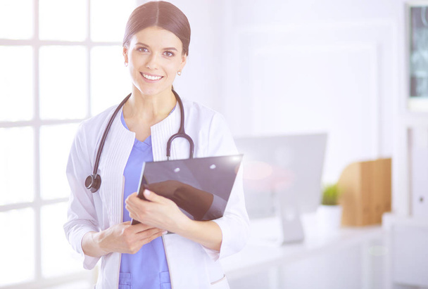 Young smiling female doctor with stethoscope holding a folder at a hospitals consulting room - Zdjęcie, obraz