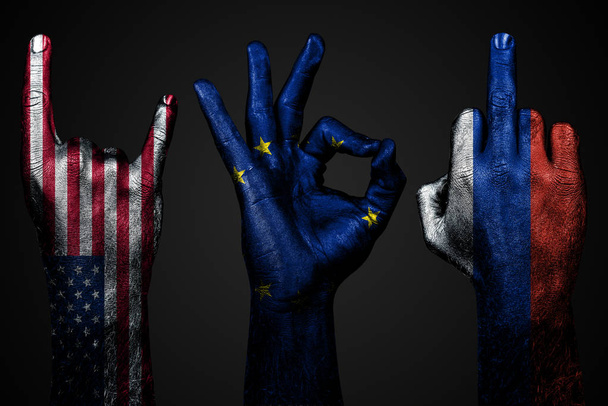 um conjunto de três mãos com uma bandeira pintada dos EUA, a União Europeia ea Rússia mostram dedo médio, cabra e Ok, um sinal de agressão, protesto e aprovação em um fundo escuro
. - Foto, Imagem