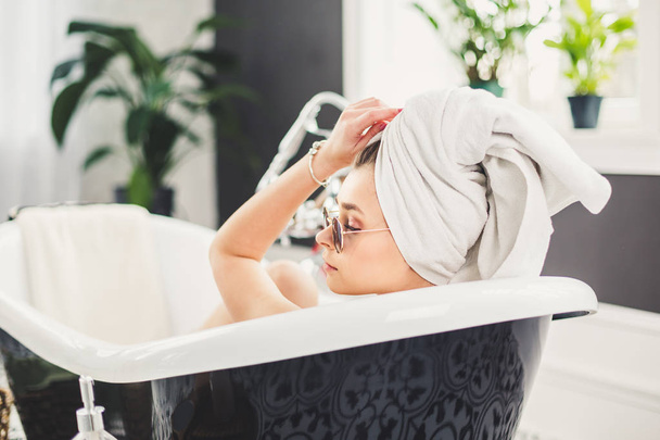 Nuori valkoihoinen nainen, jolla on pyyhe päässä ja aurinkolasit, saa kylpylähoitoa kauneushoitolassa sisätilojen huoneessa. Rentoutuminen kylpyhuoneessa liinavaatteet. Kehonhoidon ja rentoutumisen käsite
 - Valokuva, kuva