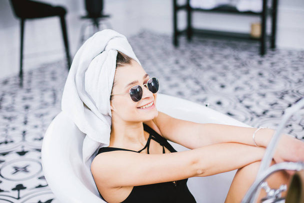 Jonge Kaukasische vrouw met handdoek op hoofd en zonnebril het krijgen van spabehandeling in een schoonheidssalon, binnen een kamer aan de binnenkant. Ontspannen in de badkamer in linnen. Het concept van lichaamsverzorging en ontspanning - Foto, afbeelding