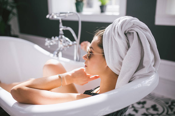 Young caucasian woman with towel on head and sunglasses getting spa treatment in a beauty salon, inside an interior room. Relaxing in the bathroom in linen. The concept of body care and relaxation - Photo, Image