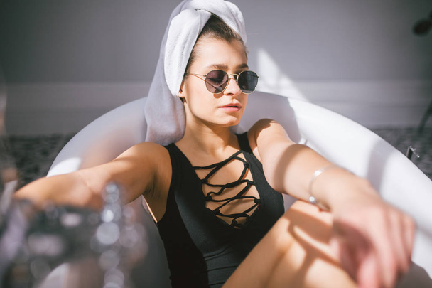 junge kaukasische Frau mit Handtuch auf dem Kopf und Sonnenbrille, die sich in einem Schönheitssalon in einem Innenraum einer Wellnessbehandlung unterzieht. Entspannung im Badezimmer in Bettwäsche. das Konzept der Körperpflege und Entspannung - Foto, Bild