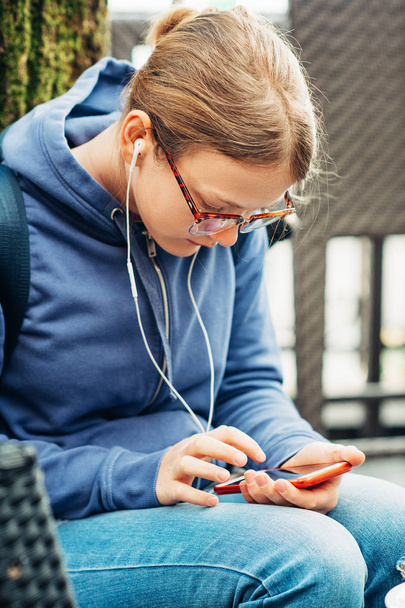 Retrato ao ar livre da jovem menina pré-adolescente vestindo mochila e óculos, ouvindo música com fones de ouvido, jogando com smartphone
 - Foto, Imagem