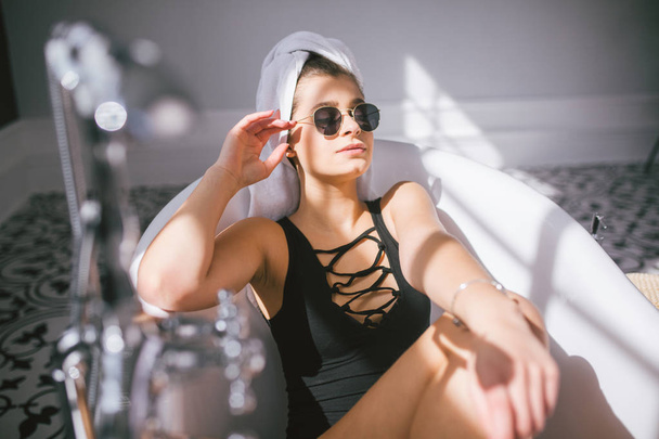 Jeune femme caucasienne avec serviette sur la tête et lunettes de soleil obtenir un traitement spa dans un salon de beauté, à l'intérieur d'une pièce intérieure. Détente dans la salle de bain en lin. Le concept de soin du corps et de relaxation
 - Photo, image