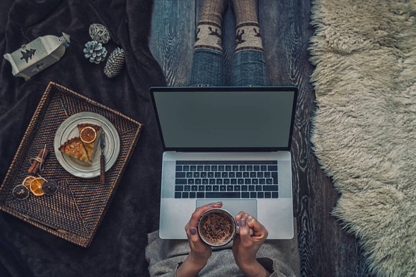 Woman in cozy home wear relaxing at home, drinking hot cacao or chocolate, using laptop. Blank screen with copy space for your text message or advertising. Christmas holidays concept. Top view. - Photo, image