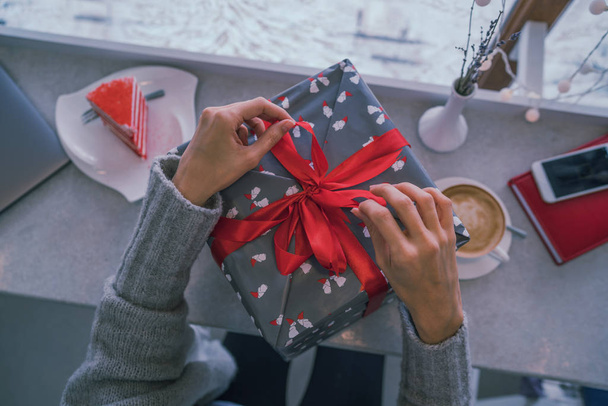 Close up shot of female hands holding a big gift wrapped with red ribbon. Big gift in the hands of a woman indoor. Shallow depth of field with focus on the big box. - Фото, изображение