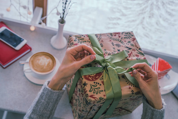 Close up shot of female hands holding a big gift wrapped with green ribbon. Big gift in the hands of a woman indoor. Shallow depth of field with focus on the big box. - Foto, afbeelding