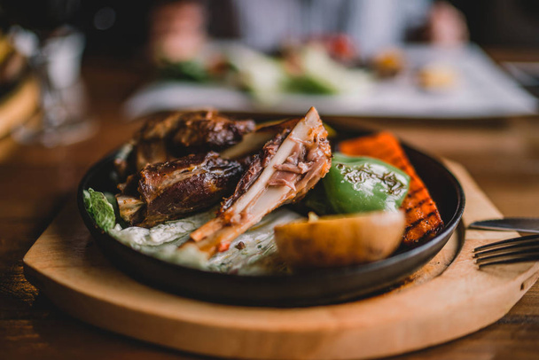 Spicy grilled lamb leg with vegetables and herbs on a roasting cast iron. Barbecue lamb with vegetables. Healthy food. Eating and leisure concept. Woman having dinner at table with food. Toned image. - Фото, зображення