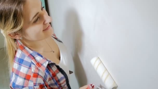 Βίντεο από όμορφη χαμογελαστή νεαρή γυναίκα ζωγραφική τοίχους στο σπίτι της με λευκή μπογιά και ρολό - Πλάνα, βίντεο