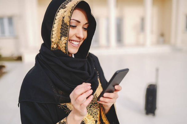 外に立ちながらスマートフォンを使って伝統的な服を着た魅力的な笑顔のイスラム教徒の女性の肖像画. - 写真・画像
