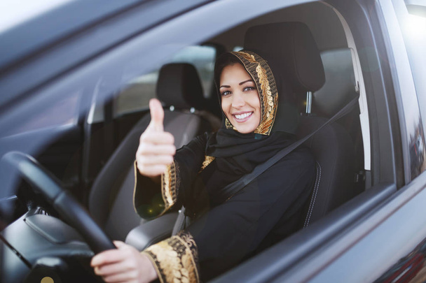 Charmante femme musulmane vêtue d'une tenue traditionnelle tenant une main sur le volant et avec un autre pouce vers le haut
. - Photo, image