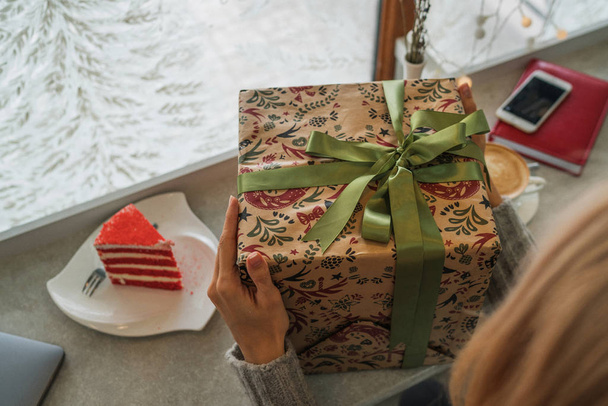 Close up shot of female hands holding a big gift wrapped with green ribbon. Big gift in the hands of a woman indoor. Shallow depth of field with focus on the big box. - Фото, изображение
