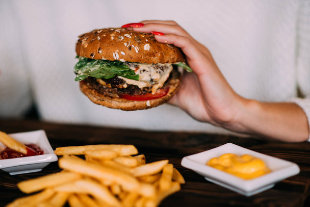 Woman holds burger in hands and potato fries on background in cafe. Hamburger made of beef, onion, tomato, lettuce, cheese and spices. Fresh burger on wooden rustic table. - Foto, Imagen