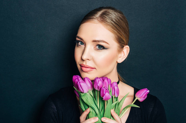 Close up portrait of beautiful young woman with dark professional make up, posing on black background, holding purple tulips - Photo, Image