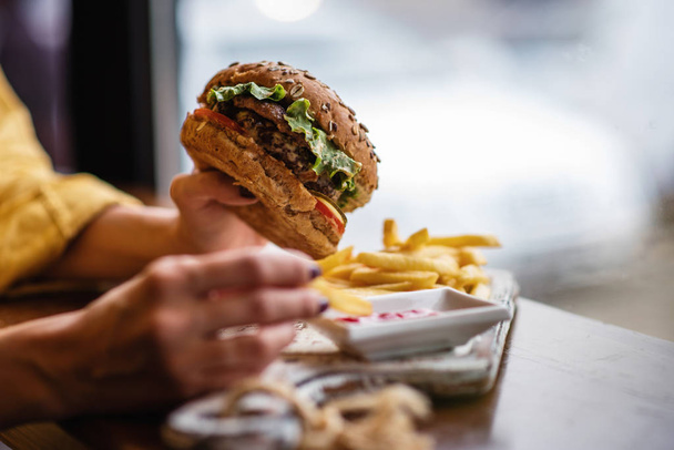 Young woman is eating fast food. Woman is eating burger while spending time in cafe. Tasty vegan burger with lettuce and beet served on cutting board. Side view with free spase for you text. - Foto, Bild