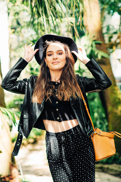 Outdoor portrait of beautiful young woman wearing black leather jacket and hat, green tropical leaves on background - 写真・画像