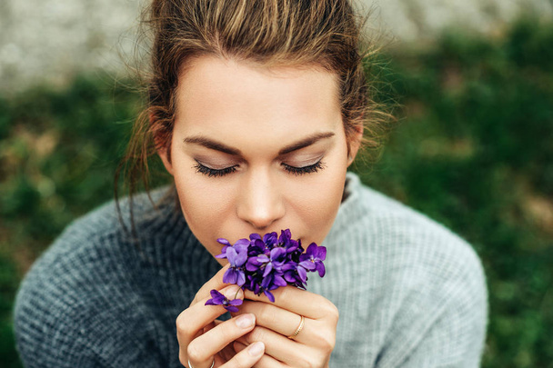 Retrato primaveral de una bonita joven con un jersey gris cálido, sosteniendo un pequeño ramo de Woolly Blue Violet (Viola sororia
) - Foto, Imagen