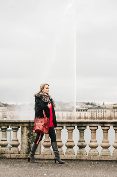 Venkovní módní portrét krásné ženy se světlými vlasy, s černým kožním kabátem, červeným pulovníkem, pózováním u jezera, jet d ' Eau fontána na pozadí, Ženeva, Švýcarsko - Fotografie, Obrázek