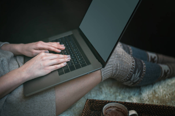 The girl sitting on a wooden floor. Laptop and coffee cup. Feet in woollen socks. The woman relaxes in cozy home. Winter and Christmas holidays concept. Selective focus. Toned image. - Fotoğraf, Görsel