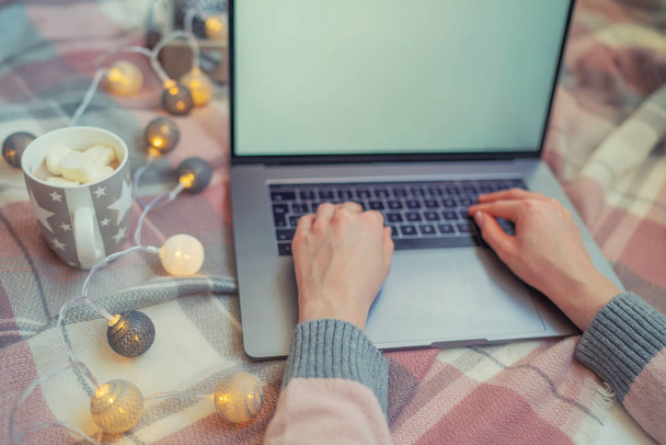 Woman's hands typing on laptop keyboard. Study and work online. Warm pink sweater, winter, home comfort and relax. Workspace with laptop, girl's hands, cake, cristmas gifts on warm plaid. - Photo, image