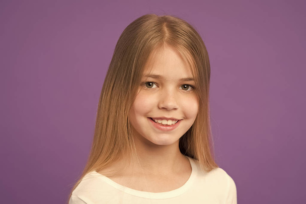 Girl on smiling face with long hair wears white shirt, violet background. Girl likes to look cute, stylish and fashionable. Kid girl with long hair looks adorable. Hairstyle and hair care concept - Fotó, kép