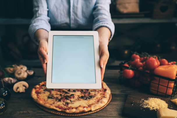 Hand holding a tablet computer with white screen. Woman hands showing empty screen of modern digital tablet. Cooking and digital everyday life online concept. Screen in focus with woman in kitchen. - Fotoğraf, Görsel