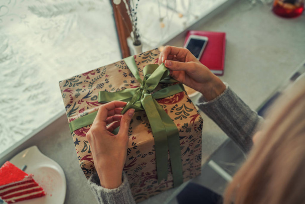 Close up shot of female hands holding a big gift wrapped with green ribbon. Big gift in the hands of a woman indoor. Shallow depth of field with focus on the big box. - Φωτογραφία, εικόνα